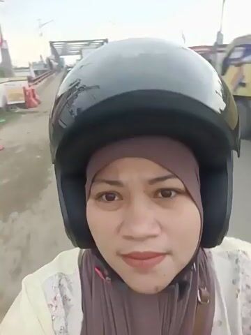 A medium shot captures a woman wearing a black helmet and a brown hijab as she rides a motorcycle. Her face is illuminated by the overhead sunlight, and she has subtle makeup on, including lipstick and eyeliner. She is looking directly at the camera with a neutral expression. The background shows a street scene with several vehicles, including trucks and motorcycles. The road appears to be paved, and there are some traffic signs and structures visible in the distance. The overall lighting suggests it is daytime, possibly late afternoon, with a slightly hazy sky.