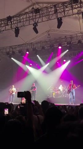 A band of four men performs on a brightly lit stage to a crowd of fans holding up their phones to record the show. Three of the band members play guitars while the fourth plays drums. The stage is equipped with several white and pink spotlights that illuminate the performers and the audience. The backdrop is a metal grid supporting the lighting equipment. The venue seems to be an indoor concert hall, filled with enthusiastic concertgoers.