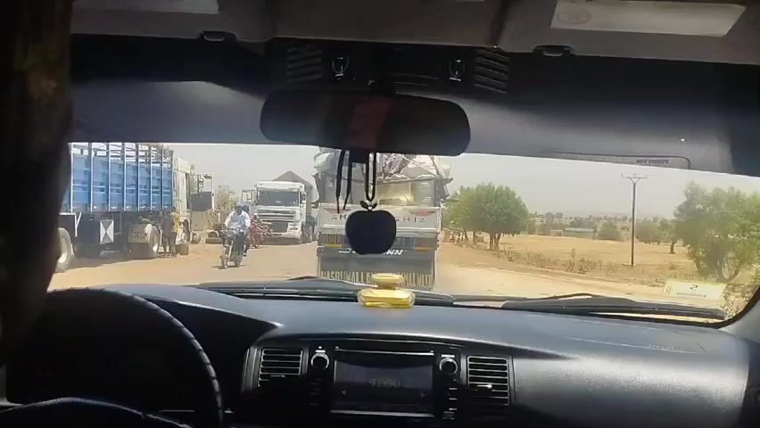 The video is shot from inside a car stopped at a dusty intersection. In front of the car, there are several large trucks. One truck is labeled "MITSUBISHI" and has multiple people sitting on top of its load of what appear to be bags of grain or similar material. There are also several people on motorcycles near the trucks. The environment is dry and rural, with sparse vegetation and utility poles visible in the background. The car's dashboard is visible, including the steering wheel, radio, and rearview mirror with a hanging apple-shaped ornament. The overall atmosphere is somewhat chaotic, likely due to the congestion caused by the trucks.