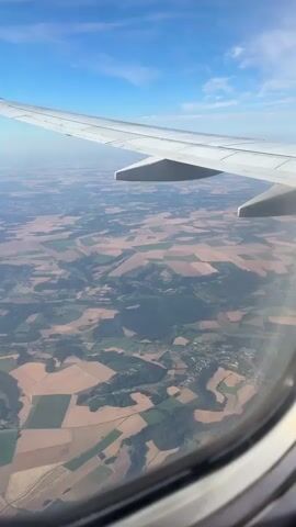 The video shows a view from an airplane window looking down at a patchwork of agricultural fields and a small town. The airplane wing is visible in the upper left quadrant of the frame, and the sky with scattered clouds is in the upper right. The landscape below is a mix of brown and green fields, indicating different crops or stages of harvest. A small village or town is visible nestled among the fields, with houses and buildings. The perspective suggests the plane is at a cruising altitude. The lighting indicates it is daytime, likely mid-morning or afternoon, given the clear blue sky and the way the sunlight illuminates the landscape. There are no people or any other discernible objects or actions occurring within the frame besides the view itself.