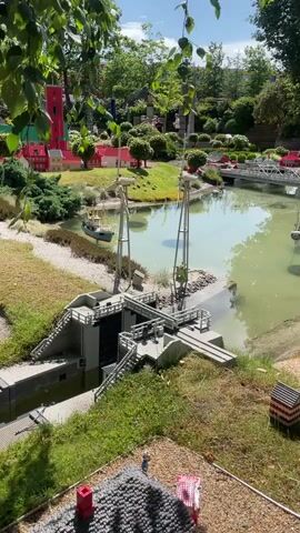 The video shows a miniature model of a town with a canal and a lock system. Two large cranes are positioned near the lock gates. A small boat is visible in the canal. In the background, there are small houses with red roofs, trees, and green landscaping. The scene is bathed in sunlight, and there are some people in the distance. The camera angle is slightly elevated, looking down on the scene.