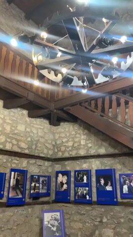 The video shows an interior scene with a stone wall and a wooden staircase. A large, industrial-style chandelier hangs from the ceiling, with multiple lights illuminating the area. Below the chandelier, a series of framed photographs are displayed on the stone wall. The photographs appear to be portraits of people, possibly in formal attire. The overall atmosphere is rustic and somewhat historical.