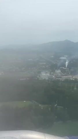 This is an aerial view from an airplane window, looking down at a hazy landscape. In the foreground, a portion of the airplane's wing is visible. The middle ground shows a mix of green trees and a large industrial complex with several buildings and visible smoke plumes rising from stacks. Further in the background, there are more buildings and distant, mist-covered hills or mountains. The overall atmosphere is foggy or overcast, obscuring finer details of the scenery.