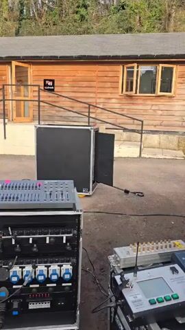 The video shows a fire effect being tested in an outdoor setting.  In front of a small, light brown wooden building with a gray roof, there is a black equipment case that emits multiple bursts of flames upwards. The fire bursts are accompanied by a whirring sound. In the foreground are equipment racks containing a sound mixing board and other electronic components connected by cables. Trees are visible in the background behind the building.