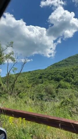 The video shows a scenic view of a hillside with lush green trees under a partly cloudy blue sky. The camera appears to be moving, likely from a vehicle, as roadside elements such as a guardrail and trees pass by. A blue sign with the name "Cetinici" is briefly visible. Power lines also appear overhead at one point.