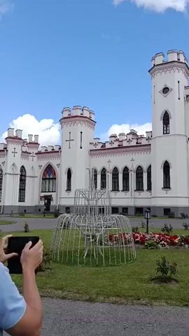 The video shows a person from behind, wearing a light blue polo shirt, walking towards a white castle with pink trim. The castle has multiple towers and arched windows. In front of the castle is a circular metal structure, possibly an unfinished fountain, surrounded by a lawn and flowerbeds with red and pink roses. The sky is blue with scattered clouds. Another person, a young woman with dark hair, walks past the fountain in the background. The camera pans slightly to the right, revealing more of the castle's facade and a paved path. The overall scene is an outdoor view of a grand building and its grounds on a sunny day.