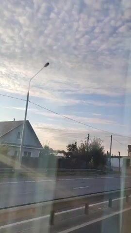 The video is taken from a vehicle moving along a road. The sky is mostly clear with scattered clouds. On the right side of the road, there are buildings, a lamppost, and a yellow car. The buildings appear to be commercial, with signs visible. There is a guardrail along the side of the road. The perspective suggests the camera is looking out a window.