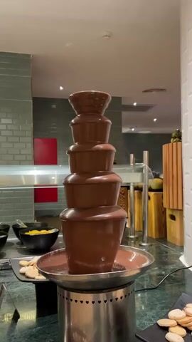 The video shows a close-up view of a chocolate fountain in a buffet setting. The fountain has multiple tiers, with the chocolate cascading down in a continuous flow. The base of the fountain is a round, stainless steel bowl filled with melted chocolate. Around the fountain are various food items, including bowls of mango pieces and plates of cookies, likely for dipping into the chocolate. The fountain stands on a green marble countertop. In the background, there's a wall with grey tiles and red squares, along with wooden boxes. The lighting in the area is bright, typical of a hotel or restaurant buffet. There are no people visible in the frame.