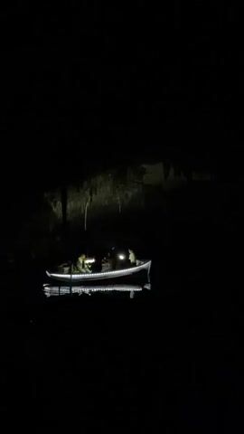 The video shows a small boat illuminated by white lights floating on water inside a dark cave. There appear to be four people in the boat. One person is pointing a flashlight towards the cave ceiling. The ceiling is partially visible, with what looks like stalactites hanging down. The water is still and reflects the lights from the boat. The overall environment is very dark.