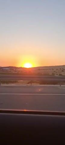 The video shows a sunrise from inside a car driving on a road. The sun is a bright yellow and orange, just above the horizon, which is lined with a low, grassy hill. A guardrail is visible beside the road. The sky is light blue. Music is playing.