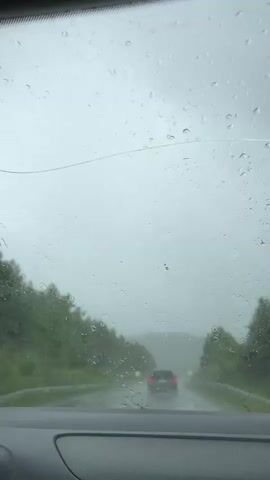 The video is filmed from inside a car, looking through the windshield. Rain is falling, and water droplets are visible on the glass. A windshield wiper is actively clearing the view. Ahead, another car is visible on a wet road, its taillights illuminated. Trees line the sides of the road, and the sky is overcast and gray. The overall impression is of driving in rainy weather on a highway.