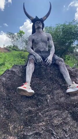A person covered in what appears to be grey paint sits on a rock formation. The person is wearing black shorts and off-white and orange sneakers. They have horns on their head and are shirtless. The person makes a gesture toward their face. Behind the person is a hillside covered in green vegetation. The sky is blue with a few scattered white clouds.