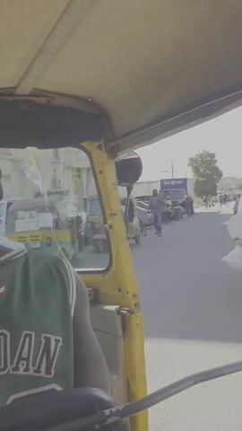 The video shows a moving view from the inside of a yellow auto rickshaw or tuk-tuk, navigating a street in a busy urban environment. People walk along the sidewalks and cross the street. Several buildings and shops line the road. One person drives a motorcycle loaded with large white bags. The driver of the auto rickshaw wears a green basketball jersey with "JORDAN" printed on the back. Other vehicles are seen parked or driving. The weather appears sunny and bright.