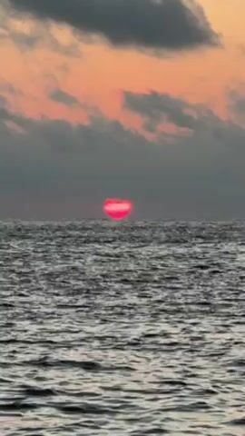The video shows a pinkish-red sun setting over the ocean. The sea is calm with small waves. The sky is a mix of orange, gray, and dark clouds. The sun appears as a flattened disc.