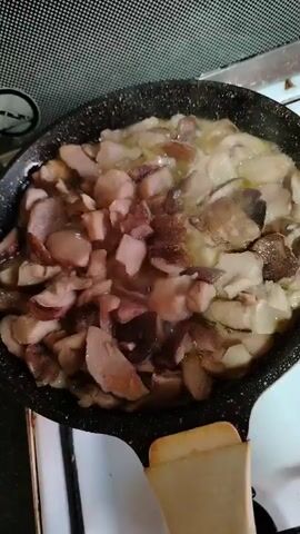 A close-up, overhead shot shows chopped mushrooms being sautéed in a black frying pan. The mushrooms are cut into irregular pieces of varying sizes and colors, ranging from light brown to dark brown. They are being cooked in a pool of hot oil or butter, which is visibly bubbling and sizzling. The pan itself has a speckled non-stick coating and a beige handle. The pan is placed on a white stovetop, and the background shows the edge of a black backsplash with a textured pattern. The lighting suggests an indoor environment, possibly a kitchen. The video appears to be a short, static recording with no apparent movement other than the cooking process within the pan.