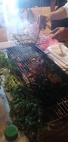 The video shows a close-up of meat grilling over hot coals. The meat is arranged on a metal grate, and a person's hands are visible, using a utensil to adjust the meat. Several bunches of green herbs are placed on the side of the grill. Smoke rises from the grill, indicating the cooking process. The scene appears to be outdoors, possibly at a street food stall, with some bottles and plastic bags visible in the background. The lighting suggests it might be evening or nighttime.