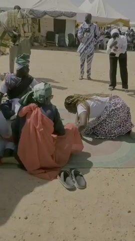 This video depicts a sandy outdoor setting with several people present. Some individuals are seated on mats, while others are kneeling and crawling on the ground, engaged in what appears to be some sort of activity or performance. A group of women dressed in matching white shirts and patterned skirts crawl forward on all fours, pushing woven baskets along the sand. A white horse stands in the background and multiple tents are set up. One woman is seen kneeling before a group of seated men. Some men are wearing traditional headwear and clothing, while others are in modern attire. Some people are seen standing and observing. The background contains buildings and vehicles, suggesting a public or semi-public location.