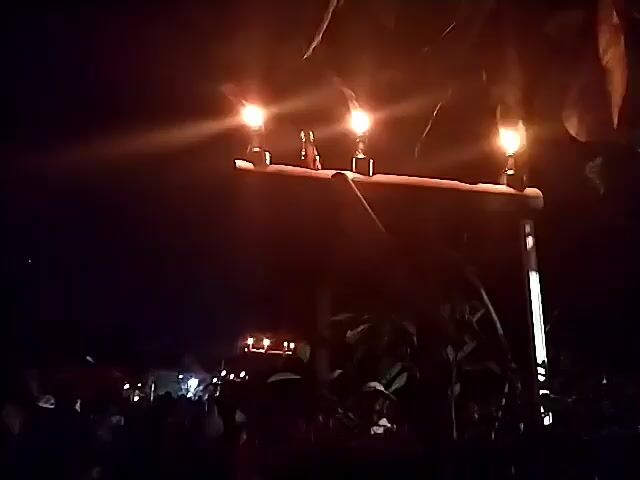 The video shows a dimly lit outdoor scene at night. In the foreground, a wooden structure holds three torches that are lit and burning, casting a warm glow. The torches appear to be held in glass containers and are mounted on a horizontal wooden beam supported by a vertical post. Beyond the torches, in the mid-ground, a crowd of people is visible, mostly in shadow. Some appear to be wearing hats. There are also glimpses of what might be tents or shelters in the background, also illuminated by lights. The overall atmosphere is dark, with the flickering flames of the torches being the primary source of light in the immediate area. The foliage of trees is also visible in the frame, particularly in the upper right corner, with leaves partially obscuring the view. The image quality is low, suggesting it was filmed with a mobile device in low light conditions.