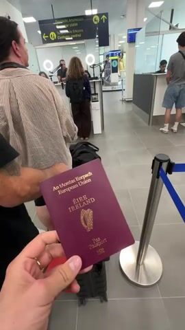 The video begins with a close-up shot of a person's hand holding an Irish passport. The passport is a deep red color and has "ÉIRE IRELAND" printed on the cover, along with the harp of Ireland and "An tAontas Eorpach European Union" text. In the background, a queue of people are waiting at what appears to be an airport or border control checkpoint. There are passport control booths with officers behind them, and signs indicating directions for "Passport Knock-off" and "Customs Control". The environment is bright and sterile, typical of an airport. The camera then slightly pans to reveal more of the queue and the surrounding area, showing luggage and other travelers. The focus remains on the passport in the foreground, with the background slightly blurred.