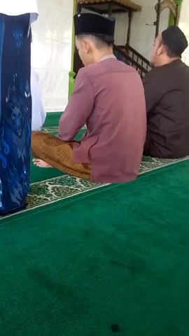The video shows a view from inside a mosque. Two men are seated on green carpeted floor. The man in the foreground is wearing a maroon colored shirt, a black cap and a sarong. The man behind him is wearing a black shirt, a black cap and a sarong. In the background, there is a man in a white shirt and a white cap, standing and holding a book. He appears to be giving a sermon or reading from the book. There are other people sitting in the background on the carpet. The mosque appears to be decorated with Islamic calligraphy on the walls.