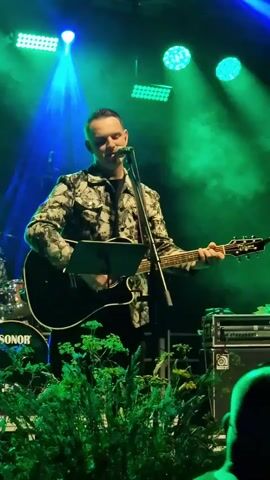 A man is playing an acoustic guitar and singing into a microphone on a stage. He is wearing a black t-shirt and a patterned jacket with a black and white floral design. The stage is lit with green and blue lights, and there is smoke or fog in the air. In the background, a drum set with "SONOR" visible on it can be seen. There are also some plants on the stage. The lower part of the frame shows the backs of people in the audience.