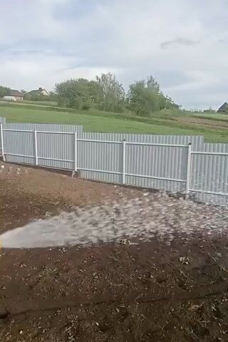 The video shows a newly laid gravel path being watered. The path runs alongside a freshly tilled garden bed with small green plants visible. A gray metal fence lines the edge of the property, with a grassy field and some houses visible in the background. The sky is overcast. The water is being sprayed onto the gravel from off-screen.