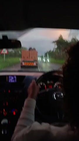 The view from inside a car is presented, focused on the driver's perspective. The car is a Mazda, apparent from the steering wheel. A person with curly hair is driving. Outside, the road is visible, with a large truck carrying a load of bricks ahead. Another car is approaching in the opposite lane. The sky is overcast with a hint of pink in the distance. The car's dashboard is illuminated with red lights. The setting appears to be a rural area with trees lining the road.