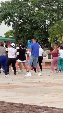 The video shows a group of people, mostly women, engaged in an outdoor dance fitness class. They are in a paved outdoor area with trees and some greenery in the background. A person in a blue shirt and denim shorts is leading the dance, while the others follow their movements. The participants are wearing athletic clothing in various colors. The overall atmosphere appears to be energetic and recreational.