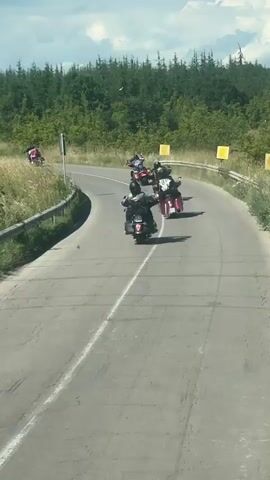 A group of motorcycles rides in formation on a winding paved road. The road is bordered by a metal guardrail on the left and tall green grass and trees on the right. Several motorcyclists are wearing helmets and riding jackets, with some also wearing backpacks. The motorcycles are various colors, including red and black, and appear to be cruisers. The sky is partly cloudy.