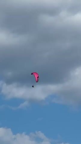 A person is paragliding in the sky. The paraglider is pink and is in the middle of the frame. The sky is blue with some clouds. There is a hill in the background.