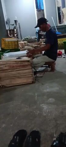A person wearing a dark blue t-shirt, light-colored pants, and a black baseball cap is kneeling on the floor. They are facing a stack of wooden crates and appear to be working with a package placed on top of them. The person's hands are visible, suggesting they are manipulating or examining the package. The environment appears to be a warehouse or storage area with concrete flooring. Several plastic crates filled with green fruits, likely mangoes, are visible in the background, along with other wooden crates and boxes. A broom leans against a wall in the distance. The lighting is dim, indicating it might be evening or indoors with artificial lighting.