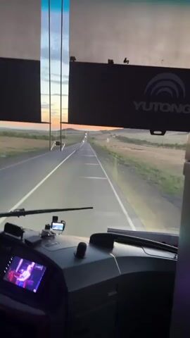 This video appears to be shot from inside a bus or similar vehicle, looking forward through the windshield. The primary subject is the road ahead, stretching into the distance under a dramatic sky. The time of day is clearly dusk or dawn, with the sky displaying hues of orange, pink, and blue, suggesting a sunset or sunrise. The landscape on either side of the road is rural, with rolling hills and dry vegetation, indicative of a steppe-like environment.

Visible objects include the dashboard of the vehicle, with a screen displaying a person on a stage, and a digital temperature reading of "24C". There is also a YUTONG logo visible on a dark panel above the windshield, which is a brand of buses. Other text includes what appears to be safety warnings or information signs.

The activity is travel, with the vehicle moving along a highway. The mood is serene, possibly even reflective, given the scenic sunset and the quiet hum of travel. The focus on the road and the distant horizon creates a sense of journey and onward movement. The presence of the screen showing a performance could imply entertainment for passengers or a live broadcast, adding a layer of modern technology to the scene.

The location context of Бастау, Kazakhstan, aligns with the visual cues of a vast, open landscape with rolling hills and a clear, expansive sky, characteristic of the Kazakh steppes.