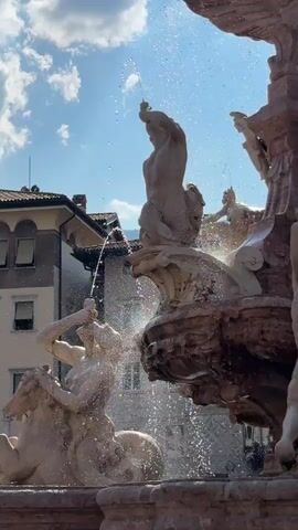 This media file captures a close-up view of a large, ornate fountain in a public square. The fountain features multiple sculpted figures, appearing to be made of stone or marble, and is actively spraying water, creating a dynamic and lively scene.

The main subjects are the elaborate sculptures. One prominent figure is a male statue in the upper portion of the frame, raising an arm as water shoots from his hand. Below him, another sculpted figure, possibly a sea creature or mythological being, is depicted with water emerging from its mouth. In the foreground, a partially visible sculpture of a horse-like creature with a human figure on its back is also prominent, with water spraying around it.

The activity taking place is the operation of the fountain, with water cascading and spraying from various points of the sculptures. This creates a visually appealing spectacle, with sunlight glinting off the water droplets, suggesting a bright and clear day.

Notable details include the architecture visible in the background, with buildings suggesting an urban European setting. The sky is a clear blue with scattered white clouds, indicating pleasant weather, likely daytime. The overall emotion conveyed is one of joy and refreshment, typical of public fountains in warm weather. There is no visible text within the media file. The location context of Trento, Italy, aligns with the architectural style and the presence of such a grand fountain.