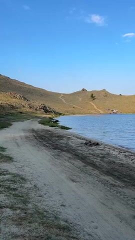A dirt road runs along the shore of a calm body of water. The water is a light blue color and reflects the sky. The road is sandy and has tire tracks on it. To the left of the road are hills covered in dry grass and rocks. There are a few scattered trees on the hills. The sky is clear and blue with a few white clouds. The scene appears to be in a rural or natural setting.