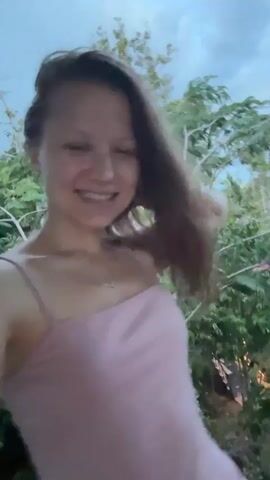 This media file features a close-up, selfie-style video of a young woman with long, dark hair and fair skin. She is smiling broadly and appears to be in good spirits. She is wearing a light pink spaghetti strap top and a delicate necklace. The background is a blur of greenery, suggesting an outdoor setting with lush foliage. The lighting appears soft, indicating it might be dusk or early morning. The woman is seen moving her hair and adjusting it behind her head, contributing to a playful and candid feel. In the blurred background, hints of structures with red roofs can be observed, along with what appear to be trees and branches. The overall impression is one of a relaxed and happy moment captured outdoors.