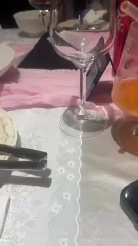The video shows a dinner table setting with multiple people. There are plates of food, glasses, cutlery, and beverages on a white tablecloth with a floral pattern. The food includes pasta, salad, and other dishes. Several people are seated around the table, some of them using their phones. The background shows a room with a window. The overall atmosphere appears to be a casual gathering or meal.