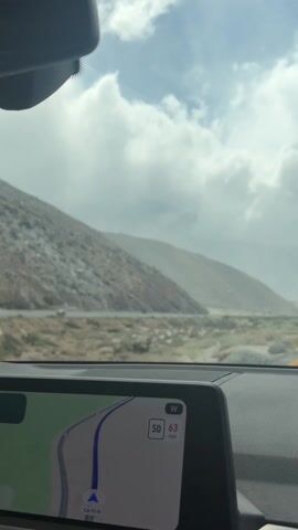 The video shows a car driving on a highway with a desert landscape and mountains in the background. The sky is overcast and cloudy. The car's navigation system is visible, showing the route and speed, which is 63 mph. There are other cars on the road.