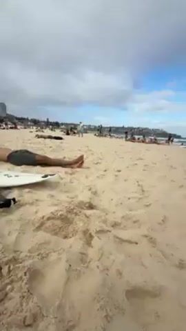 The video begins with a first-person view of a sandy beach on an overcast day with patches of blue sky. People are scattered on the beach, some near the water's edge. A shirtless person comes into frame and looks at the camera briefly before the camera angle is changed. A lifeguard stand is visible in the background. The camera is angled upwards, showing the sky and the lifeguard stand, then returns to the beach view.