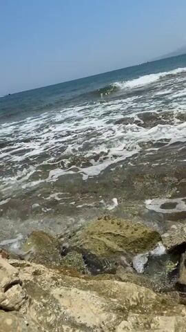 The video shows a close-up, low-angle view of the ocean waves crashing against rocks on a shore. The water is clear and turbulent, with white foam from the waves. The rocks are varied in size and color, some appearing smooth and others rough. In the distance, the ocean stretches out to the horizon under a clear blue sky, with faint landmasses visible on the far shore. The sun is shining, creating a bright and natural light on the scene. There are no people or animals visible in the video.