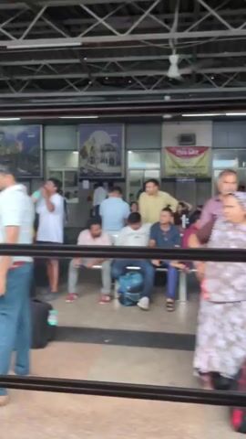 The video shows a crowded indoor area, likely a train station waiting area. People are seen sitting and standing, many with luggage. The environment appears to be a covered platform with overhead lighting and a metal support structure. Posters and signs are visible on the walls. The people are dressed in casual clothing, and there are individuals of various ages. Some people are looking at their phones, while others are talking or waiting. The overall atmosphere suggests a busy transit hub.