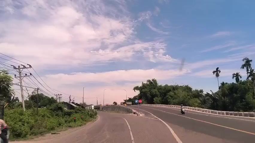 The video depicts a roadway scene under a bright, partly cloudy sky. The camera, presumably mounted on a vehicle, is approaching a Y-intersection. To the left, a narrow road leads toward a cluster of simple structures nestled amidst lush green vegetation. Electrical lines are visible overhead, strung between poles. Straight ahead and to the right, a wider road ascends onto a bridge. The bridge is bordered by a concrete barrier. Signage indicating traffic rules is present on the right side of the road. A vehicle is seen driving on the right side of the road over the bridge. Palm trees and dense foliage form a backdrop along the right side of the road.