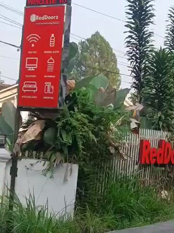 A person is riding a motorcycle down a paved road. To the left of the road is a red sign for RedDoorZ, which lists amenities like free WiFi, mineral water, television, and a bathroom. Further down the road, a cluster of red buildings with white trim are visible, adorned with red and white striped flags. Several other motorcycles are also on the road. The environment appears to be a rural or suburban area with lush greenery and trees.
