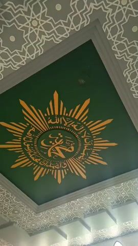 A downward-angled shot focuses on a ceiling adorned with Islamic calligraphy and geometric patterns. The central element is a large, sunburst-like emblem in gold on a dark green background. Within the emblem, Arabic script is visible. The surrounding ceiling features intricate, repeating geometric designs in a lighter color against a gray background. The edges of the ceiling are framed by white molding. Below the ceiling, in the background, a row of arched windows can be seen, letting in light. The overall impression is of a richly decorated interior, likely a mosque or similar religious space. There are no people visible in the frame.
