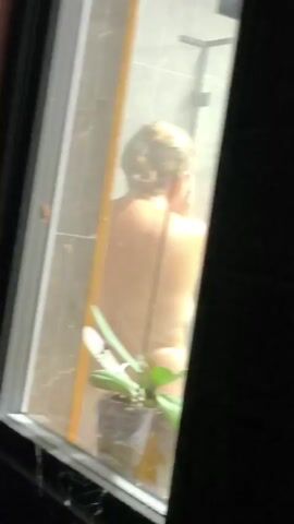 A person is showering in a bathroom visible through a glass door. The person is facing away from the camera and appears to be washing their face or hair. There is a potted plant with green leaves on the windowsill in front of the shower. A spider is visible on one of the plant's leaves. The shower has tiled walls and a showerhead.