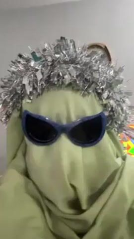 A person is covered in a light green cloth and has on a silver garland crown and blue framed sunglasses with dark lenses. Behind the person, a clock on the wall and part of a colorful TV are visible. The person remains still.