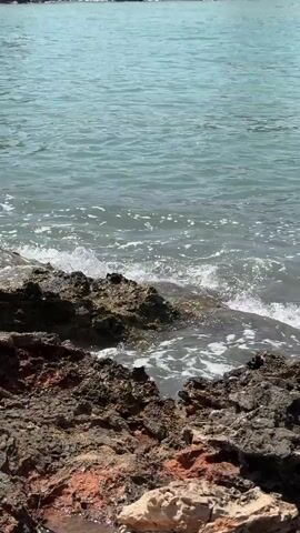 A video taken at an ocean shore shows waves crashing against a rocky coastline. The water is a clear blue-green and appears calm in the distance, but grows choppier as it nears the shore. The rocks are dark and jagged, with some reddish-brown coloration visible, suggesting a volcanic or mineral-rich composition. Sunlight glints off the water's surface. The camera is positioned at a low angle, focusing on the immediate shoreline. There are no people or man-made objects visible in the frame.