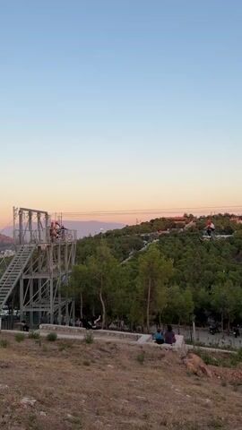 A person on a bicycle is suspended by wires above a treed area with buildings in the background. The sky is a clear gradient of blue to pale orange, indicating either sunrise or sunset. In the foreground, a metal structure with stairs and platforms is visible, with a few people on it. Further down, on a lower level, several people are seated on a stone ledge, and a few motorcycles are parked. The overall environment appears to be an outdoor recreational space.