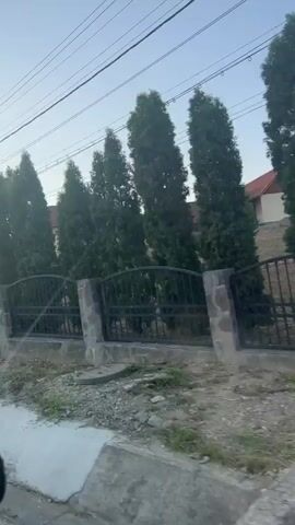 A video shows a street view, possibly from a moving vehicle. A stone and metal fence runs along the side of the road, with tall evergreen trees behind it. A house with a red roof is visible beyond the fence. The sky is clear and blue, suggesting daytime. The camera is angled towards the right, showing more of the fence and trees as the video progresses. The audio is of a song with lyrics that translate to "Here a fat diploma, a local hooligan meets us."