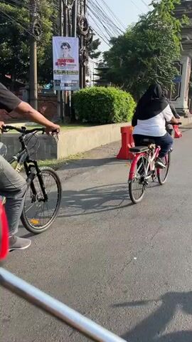 Two individuals are riding bicycles on a paved road. The first individual, in the foreground and to the left, is riding a bicycle with a red rear fender and is wearing a hijab and a white top. The second individual is further ahead and to the right, also on a bicycle, wearing a black jacket and pants. Orange traffic barriers are present along the side of the road. Trees and buildings are visible in the background. The sun is shining, casting shadows.