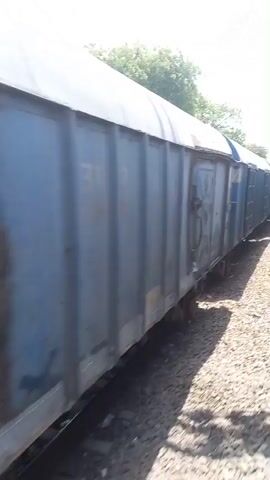 The video shows the side of a train as it moves along the tracks, passing another train traveling on the adjacent track. The train cars are various shades of blue and have a somewhat weathered appearance. The space between the tracks is filled with gravel, and there are trees and a bright, overcast sky visible in the background. The camera appears to be positioned inside a train, looking out the window.